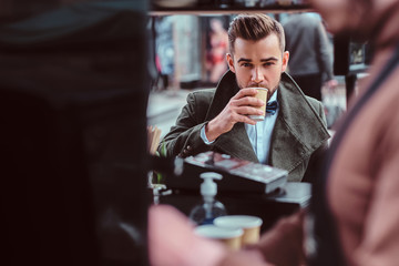 Handsome groomed man is enjoying his coffee while sitting at coffeeteria. There are working barista at frontground.