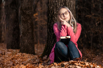 Beautiful girl in autumn forest reading a book. woman sits near a tree and holds a book