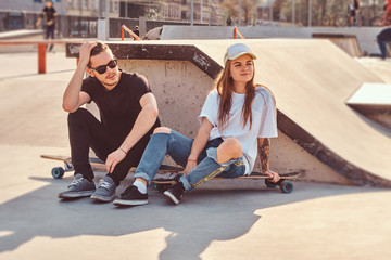 Attractive young couple is sitting at sunny skatepark with their longboards next to the ramp. - Powered by Adobe