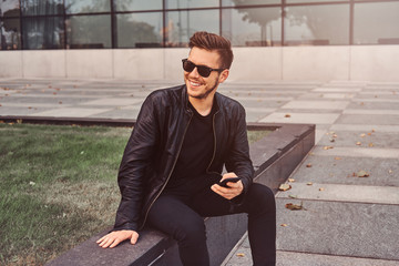 Obraz premium Smiling attractive man is chatting on mobile phone while sitting outside near building.