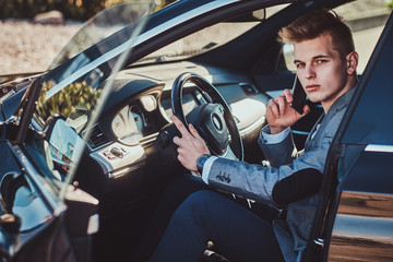 Young attractive businessman is sitting in his car at the parking while talking by mobile phone.