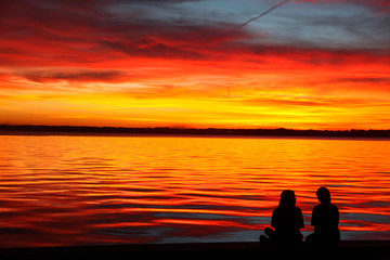 Naklejka na ściany i meble Two girls sitting with a bright orange sunset on the lake