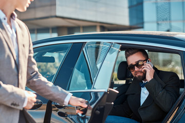 Busy nice businessman in sunglasses is talking by smartphone while his elegant assistant is opening door for him.