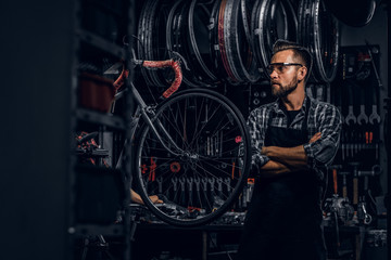 Plakat Smiling bearded man in glasses is standing near fixed bicycle at his own workshop.