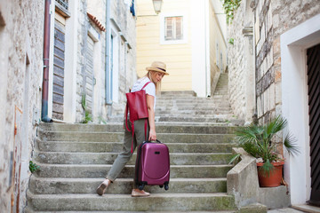 Girl traveler holds heavy suitcase on high stairs. Woman tourist carries luggage at old town street...