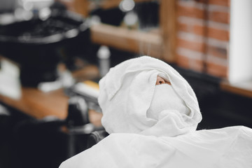 Fototapeta na wymiar Man sits in barbershop chair and steam his face with hot towel in front of royal beard shaving razor