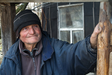 An old man in sloppy clothes stands on the threshold of his own ruined house and looks into the distance