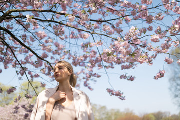 Beautiful blonde young woman in Sakura Cherry Blossom park in Spring enjoying nature and free time during her traveling tourist free time - Wearing white pants and t-shirt with a beige jacket