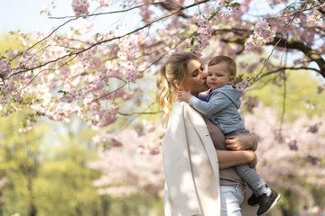 Young mother mom holding her little baby son boy child under blossoming SAKURA Cherry trees with falling pink petals and beautiful flowers