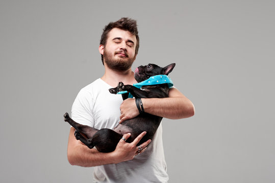 Hipster Bearded guy holding and hugging a nice French Bulldog dog in his arms with love and playing with him, against a white background