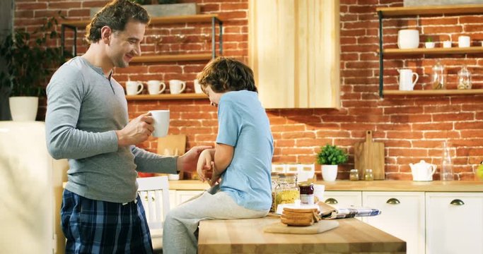 Cheerful Caucasian father in the pajama drinking coffe in the morning and tickling little son in the pajama during breakfast.