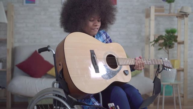 disabled african american woman with an afro hairstyle in a wheelchair plays acoustic guitar close up