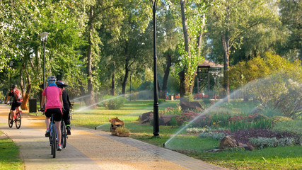 Cyclists riding bicycles in city park. Sprinkler showering lawn in summer park. Beautiful sunny day.
