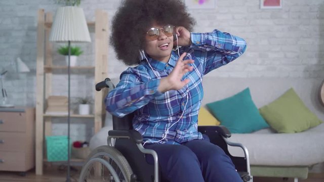 cheerful disabled african woman with an afro hairstyle in a wheelchair in headphones listening to music and singing
