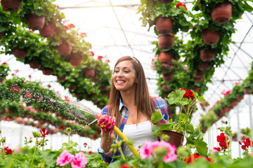 Beautiful smiling woman florist enjoying her job and watering plants with love in flower's greenhouse. Female worker taking care of plants. Water splashing out of hose, drops flying over flowers.