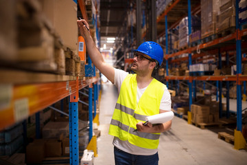 Warehouse worker checking inventory in large distribution warehouse. Industrial worker holding checklist in factory storage area.