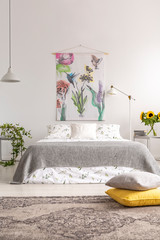 Grey and yellow pillows placed on carpet in real photo of white bedroom interior with material painting, king-size bed with floral bedding, fresh sunflowers and lamps