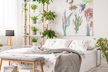 Grey blanket thrown on double bed with floral bedclothes standing in white room interior with painting, many fresh plants on wooden rack in the real photo