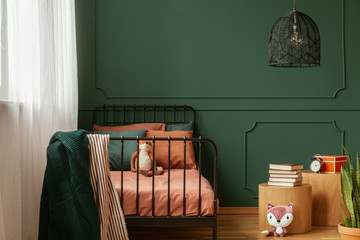 Real photo of a cute, green and orange bedroom interior for a kid with plush fox toys, molding on green wall and books on bedside table