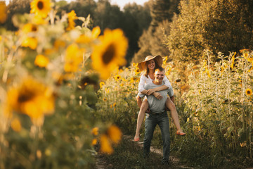 Beautiful couple in love is embracing, looking at the sunset over a field of sunflowers. Girl in a hat and white summer dress, a man in jeans and a tshirt. Summer vacation concept, recreation in the - obrazy, fototapety, plakaty