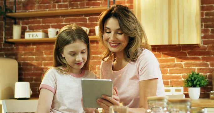 Pretty smiled Caucasian mother sitting in the cozy kitchen with a little cute daughter and showing her something on the tablet computer in the morning.
