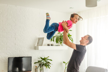 I can fly. Happy dad is lifting up his little daughter while standing in living room. Dreamful girl is wearing fairy wings and laughing