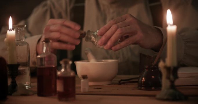A man skilled in apothecary with his bottled medicinal concoctions on the table where he is sitting working with a mortar and pestle.