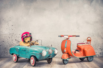 Retro Teddy Bear toy racer in old helmet hat with goggles driving antique rusty mint blue pedal car from 60s and orange kids scooter trike front concrete wall background. Vintage style filtered