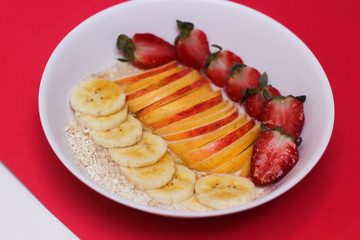 Oat Flakes with sliced bananas and honey, apples and strawberries in bowl on red background.Top view.