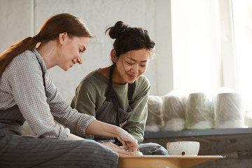 Content positive young multi-ethnic women in aprons sitting at pottery wheel and creating clay pot together