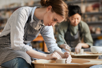Serious concentrated young woman sitting at pottery wheel and adjusting edges of clay vessel while making pot in workshop