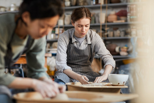 Serious attractive young female potter in stripped apron concentrated on work sitting at pottery wheel and shaping clay with hands in workshop