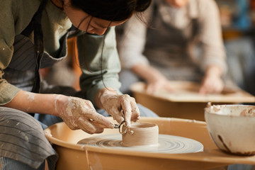 Close-up of concentrated Asian craftswoman in apron sitting at pottery wheel and using craft tool while shaping wet clay vessel - obrazy, fototapety, plakaty