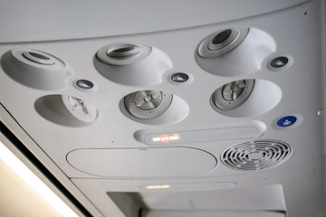Air conditioning vents, lights and buttons on ceiling of airplane above passenger seat, no smoking sign is lit