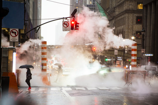 A woman with an umbrella and red high heels shoes is crossing the 42nd street in Manhattan. Cars and steam coming out from from the manholes in the background. New York City, Usa.