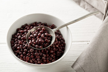 Raw, uncooked, dried adzuki (red mung) beans in white bowl with metal spoon on white wood table background