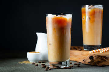 Ice coffee in a tall glass with cream poured over, ice cubes and beans on a old rustic wooden...
