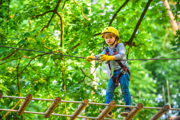 Naklejka na ściany i meble Kids boy adventure and travel. Adventure climbing high wire park. Early childhood development. Cute child in climbing safety equipment in a tree house or in a rope park climbs the rope