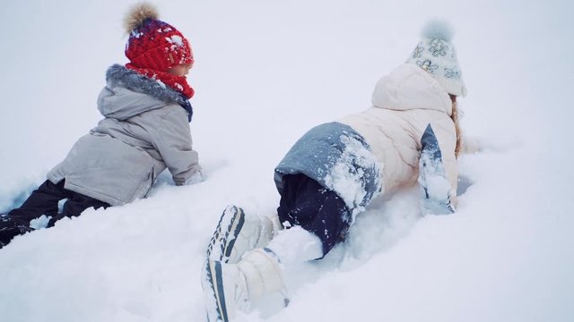 Back view of two kids crawling on snow on the white background in winter. Happy children are lying in soft snow outdoors. Close- up. Slow motion.