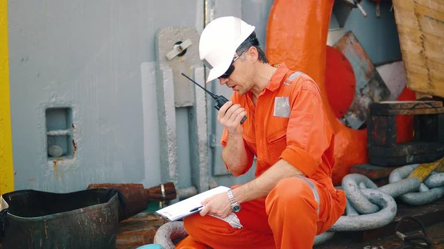 Marine chief officer or chief mate on deck of ship or vessel. He fills up ahts vessel checklist. Ship routine paperwork. He holds VHF walkie-talkie radio in hands.