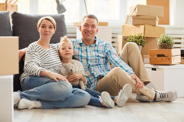 Smiling modern young family in sneakers and casual clothing sitting on floor and hugging each other while looking at camera in new flat