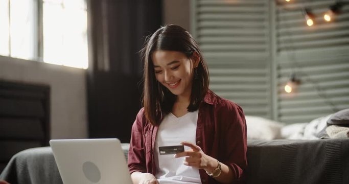 Pretty student is sitting in her bedroom with laptop on knees during online shopping. Panasian girl keying in a credit card and happily smiling 4k