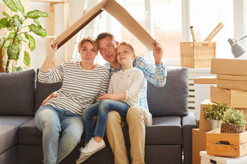 Portrait of content young family in casual clothing sitting on gray sofa and holding cardboard roof...