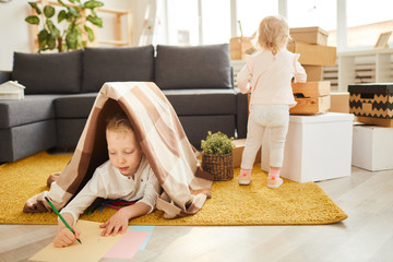 Children getting settled in new apartment: concentrated little boy lying under plaid on carpet and drawing picture while his younger sister unpacking boxes