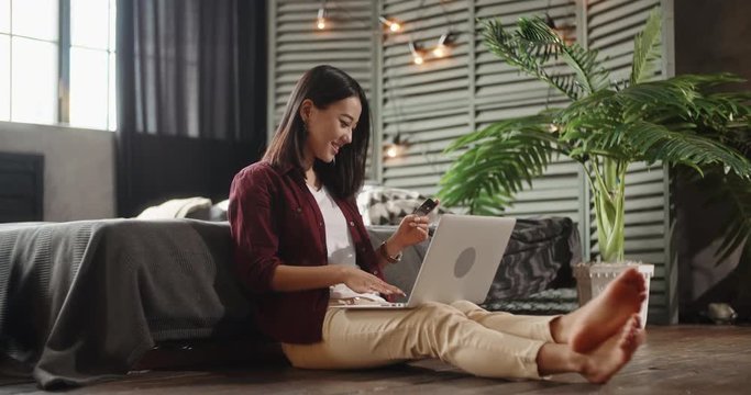 Pretty student is sitting in her bedroom with laptop on knees during online shopping. Panasian girl keying in a credit card and happily smiling 4k