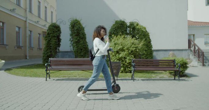TRACKING attractive African American black female walking in the street with an electric scooter, having a soft drink. 4K UHD 60 FPS SLOW MOTION