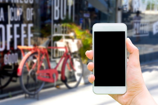Mockup image of hand holding white cellphone with blank black screen that taking pictures the vintage bicycle in front of the coffee shop is blurred on background.