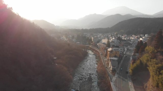 Drone flight, over small Japanese town of Nikko, misty background during golden hour with rays shining showing silhouettes of mountains in the background and a river running through the middle. 4K