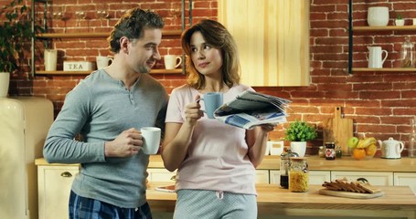 Caucasian handsome husband drinking morning coffee or tea in the morning and standing beside his wife who reading a newspaper and sipping coffee in the kitchen.