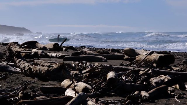 early morning winter California beach scene with large pile of driftwood in foreground and surfer waiting for opportunity to paddle out in the background as surf rolls in
